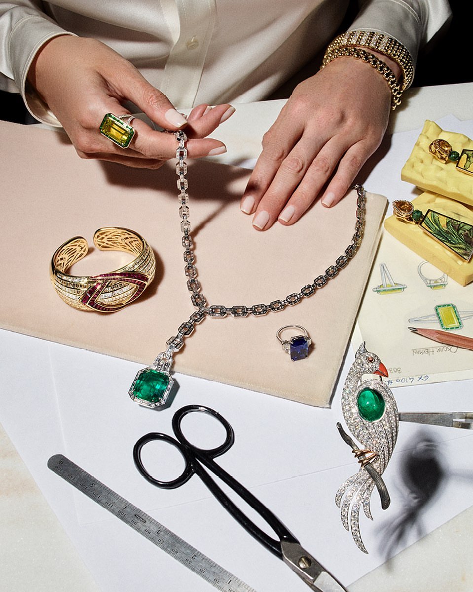 Overhead shot of hands holding diamond chain among rings, bracelets, and jewelry sketches on a white surface for Visie creative platform.