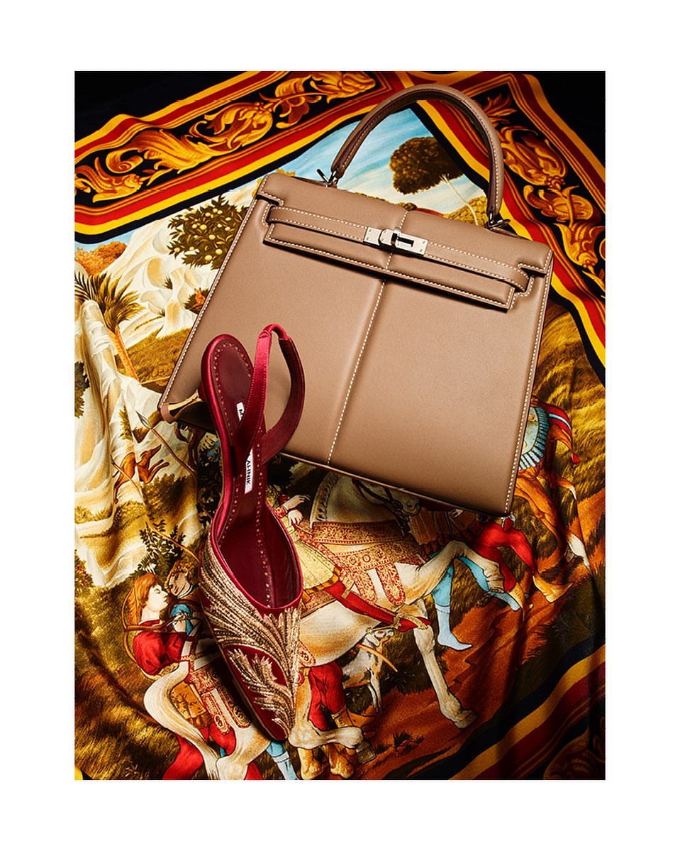 Still life shot of a beige leather handbag and a single red satin mule with gold embroidery resting on a patterned silk scarf.