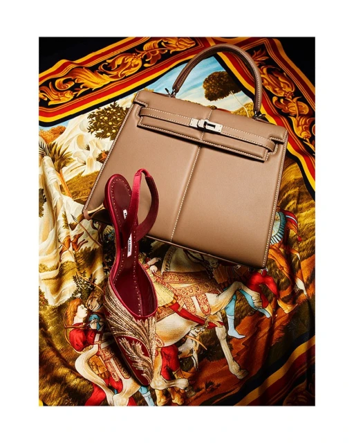 Still life shot of a beige leather handbag and a single red satin mule with gold embroidery resting on a patterned silk scarf.