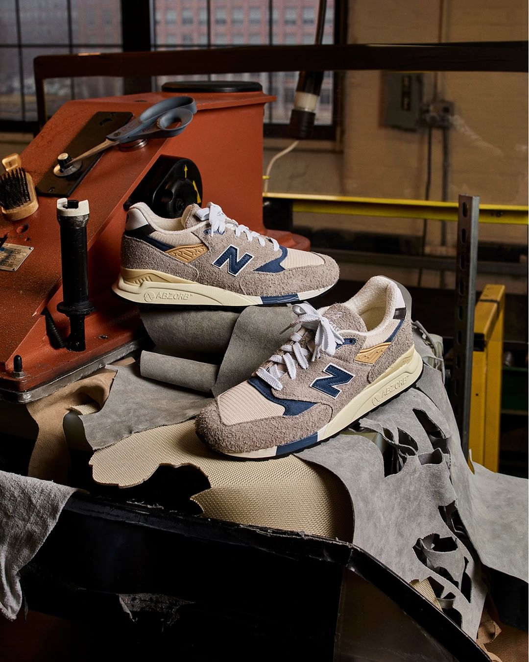 Close-up of two New Balance 998 sneakers displayed on top of leather scraps and set against a rustic industrial backdrop.