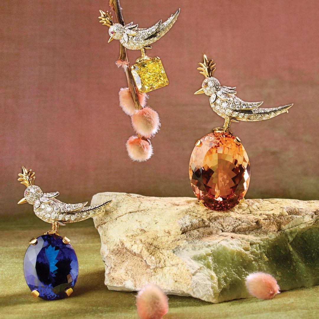 Three bejeweled birds perched on gemstones and a branch, presented against a soft pink backdrop in a still life composition.