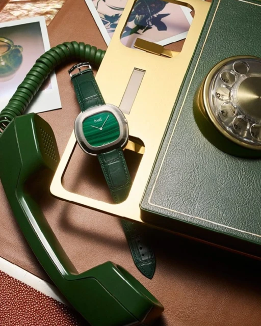 Luxury wristwatch with a malachite face and green leather strap displayed on a brown surface next to a retro green rotary telephone.