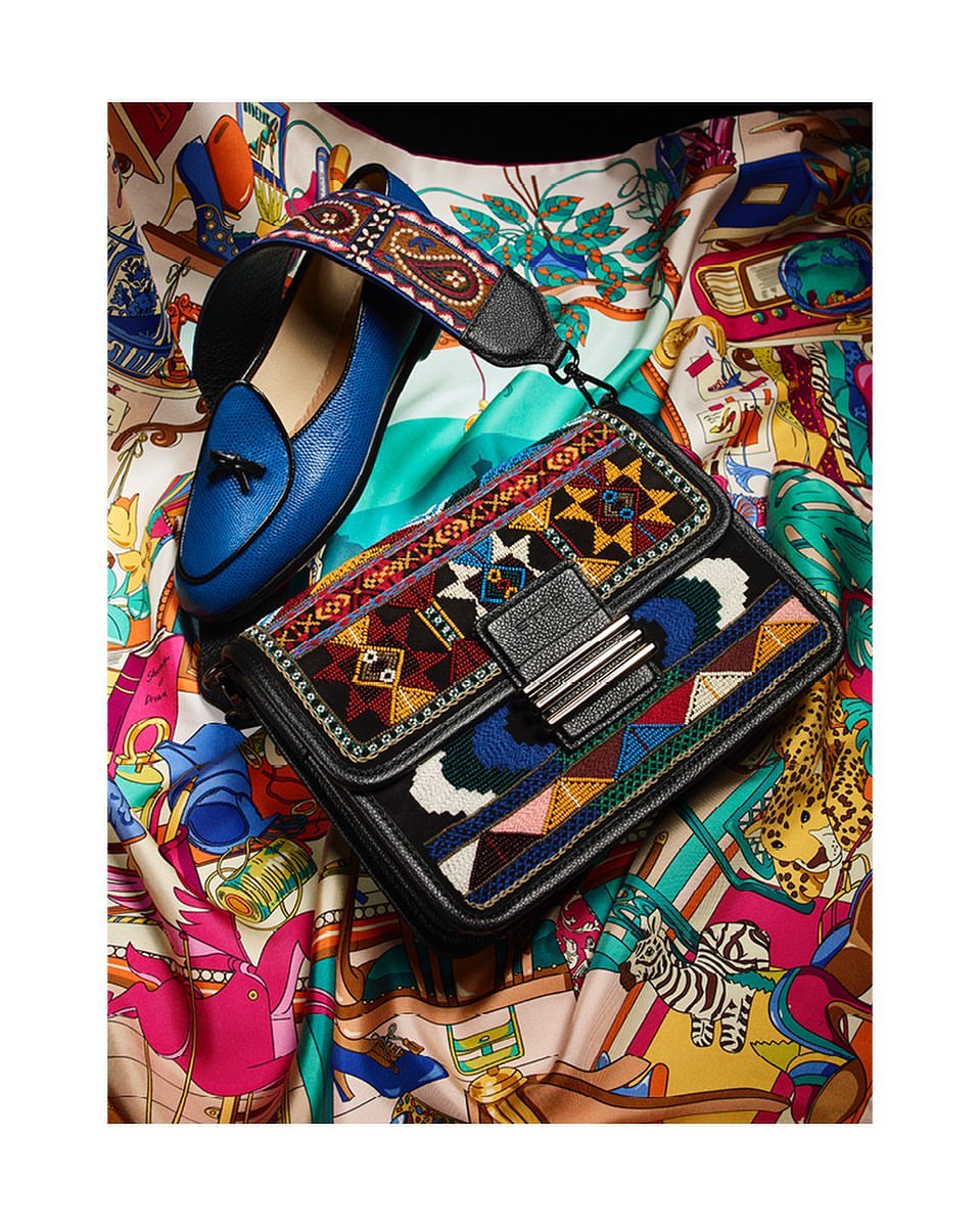 Close-up of a black leather purse with colorful embroidery next to a blue loafer on a patterned textile background.