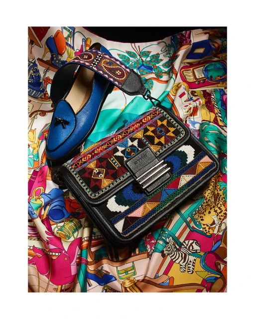 Close-up of a black leather purse with colorful embroidery next to a blue loafer on a patterned textile background.