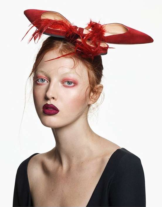 Fashion portrait of a red-haired woman wearing red lipstick, with a pair of red high heels with feathers on her head, on white backdrop.