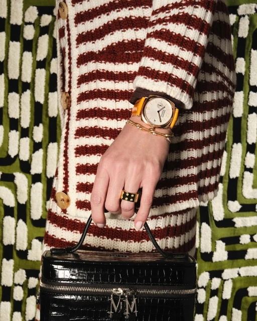 Woman showcases a gold wristwatch with an orange strap and a ring, holding a textured black purse, with patterned backdrop.