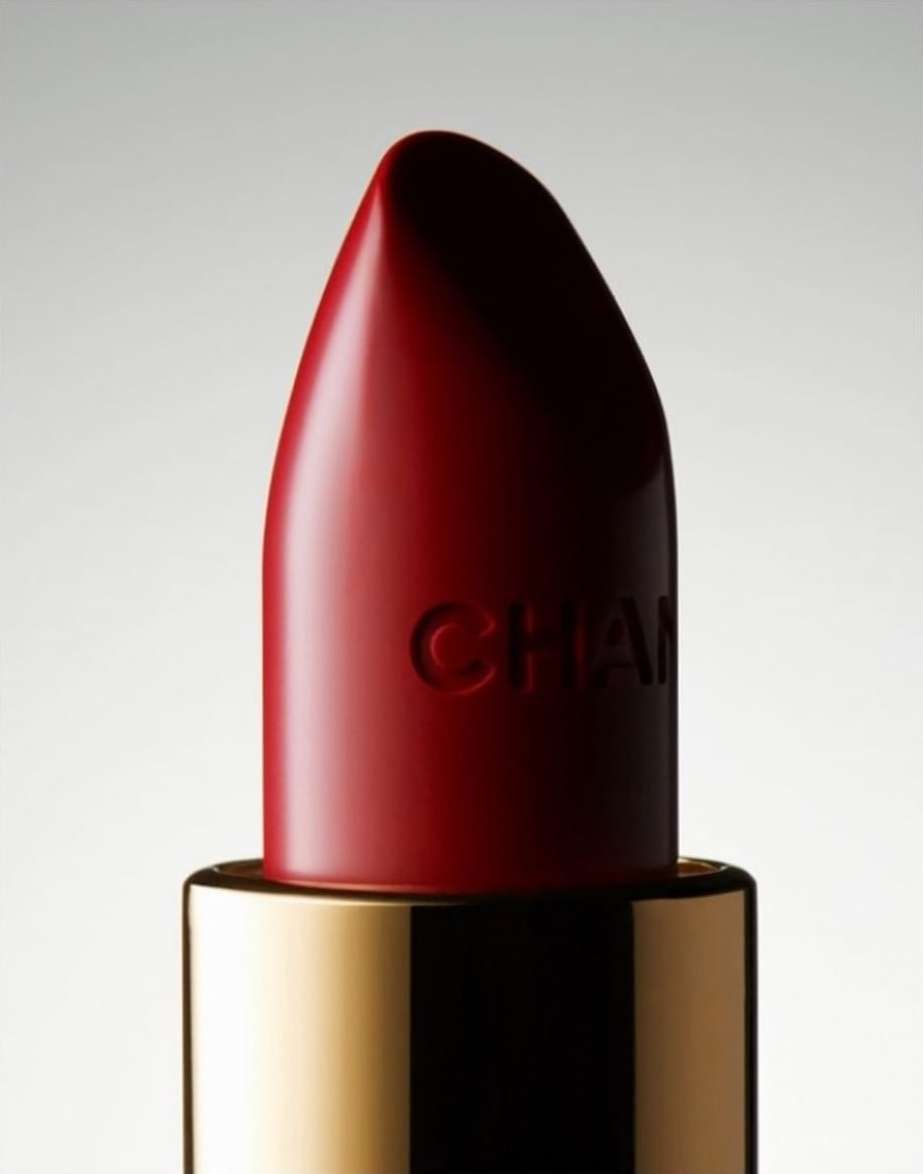 Macro shot of a red Chanel lipstick with gold base against a soft gradient background in a beauty or fashion advertising setting.