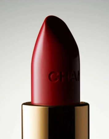 Macro shot of a red Chanel lipstick with gold base against a soft gradient background in a beauty or fashion advertising setting.