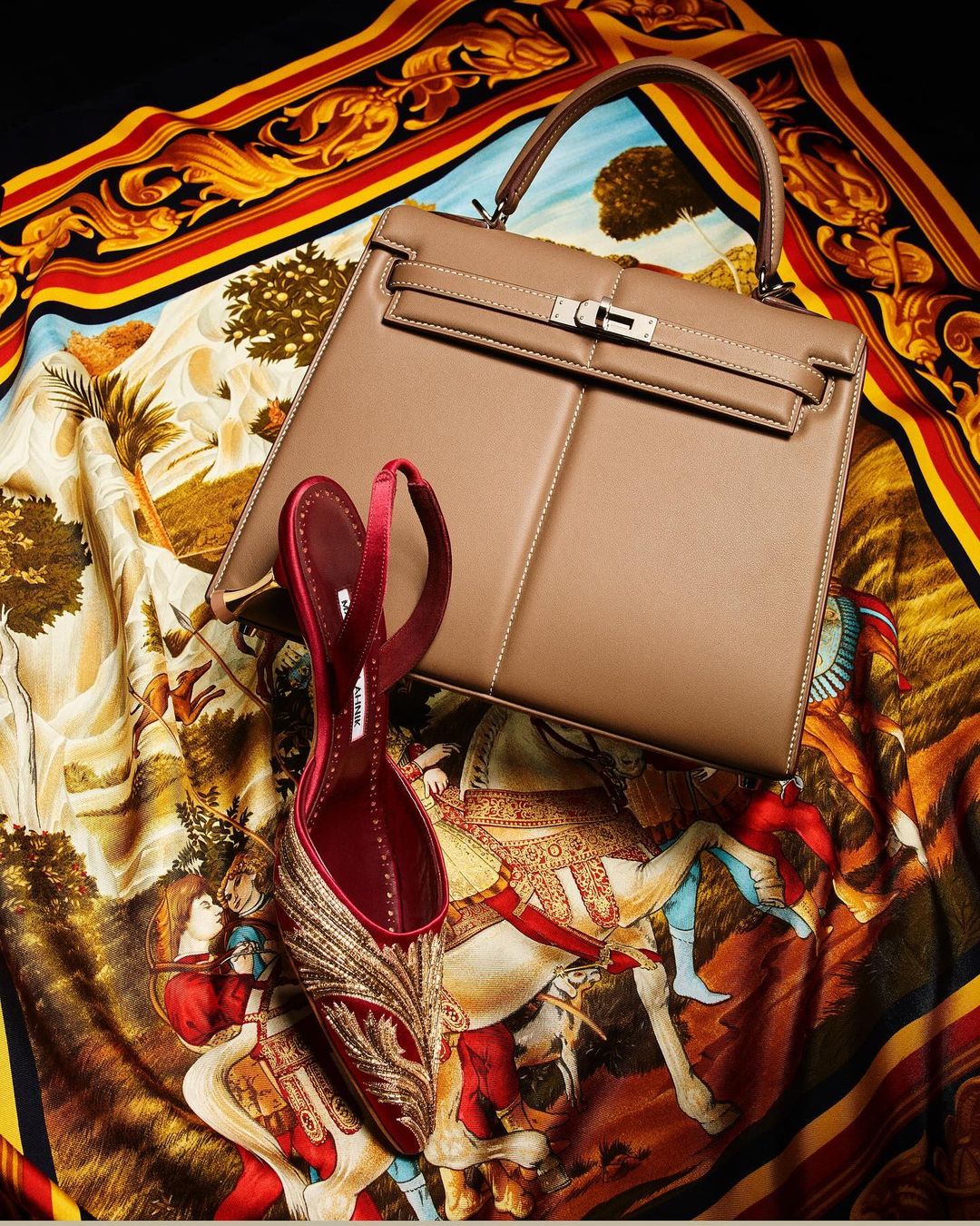 Beige leather handbag and red satin heel shoe displayed on a patterned scarf with red, yellow, and brown tones against a black background.