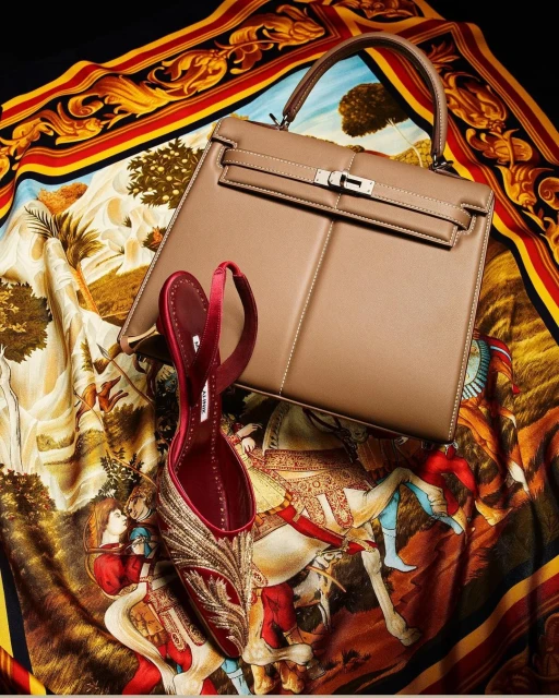Beige leather handbag and red satin heel shoe displayed on a patterned scarf with red, yellow, and brown tones against a black background.