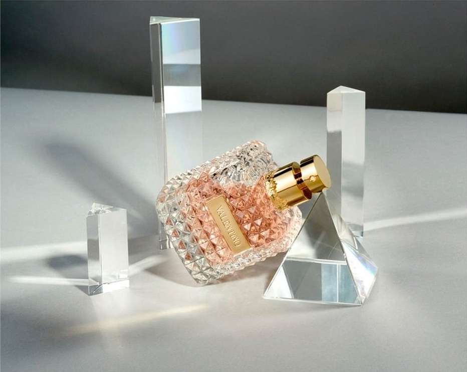 Valentino perfume bottle on a white surface surrounded by clear geometric shapes against a grey backdrop, studio lighting, luxury product.