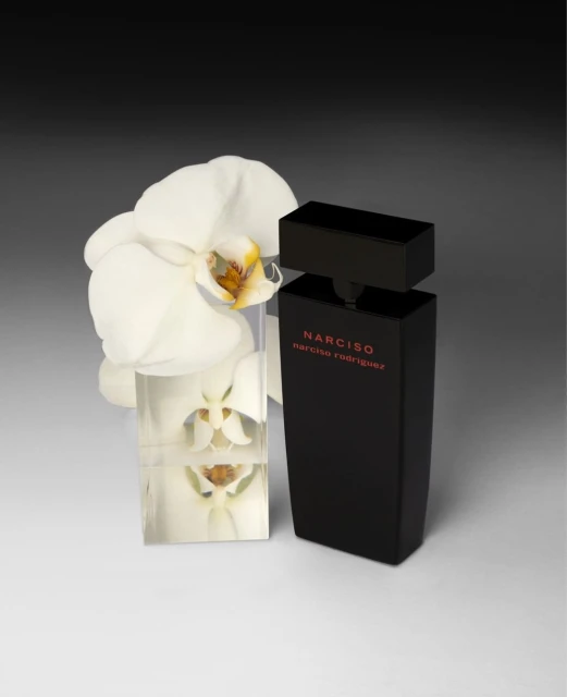 Close-up studio shot of Narciso Rodriguez perfume bottle, positioned with a white orchid on a light grey surface against dark grey gradient.