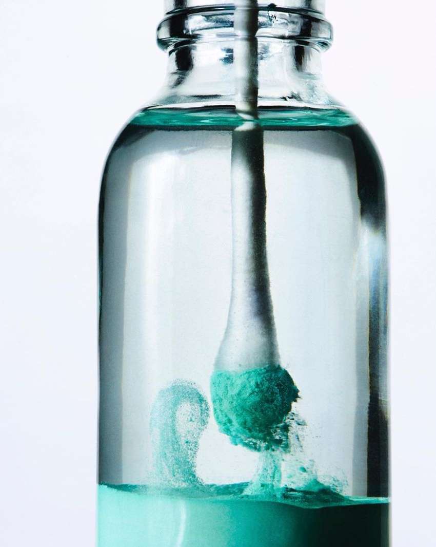 Close-up of a glass bottle with clear liquid and a cotton swab immersed in a green-tinted substance, creating a swirling effect.