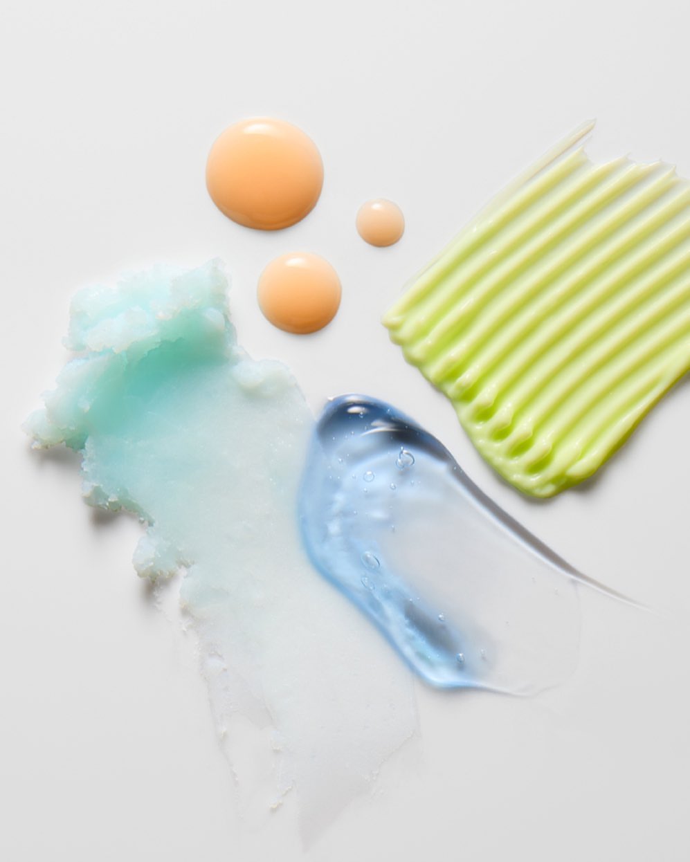 Cosmetic swatches of skincare products in orange, blue, and green tones are smeared on a white surface creating an abstract design.