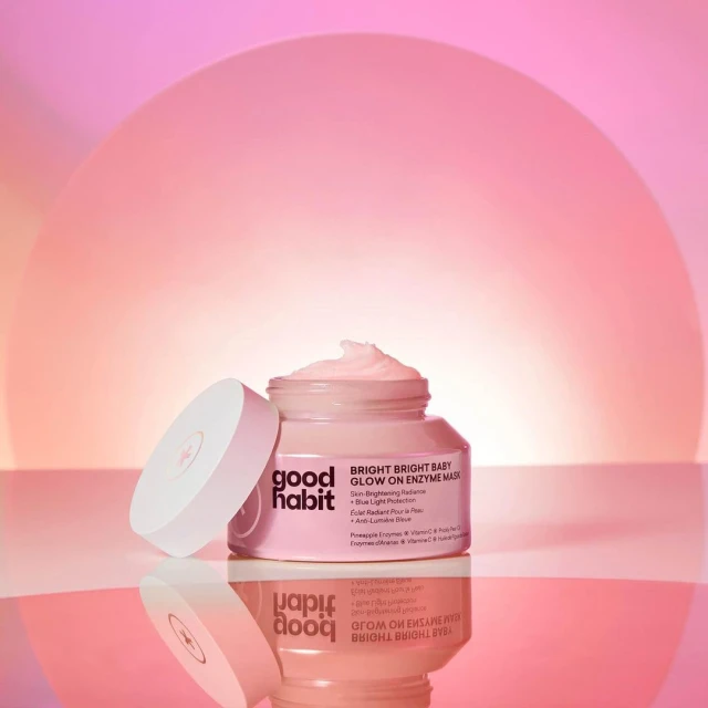 Close-up shot of a pink 'Good Habit' enzyme mask in a glass jar with a white lid, product description visible, against a gradient pink background.