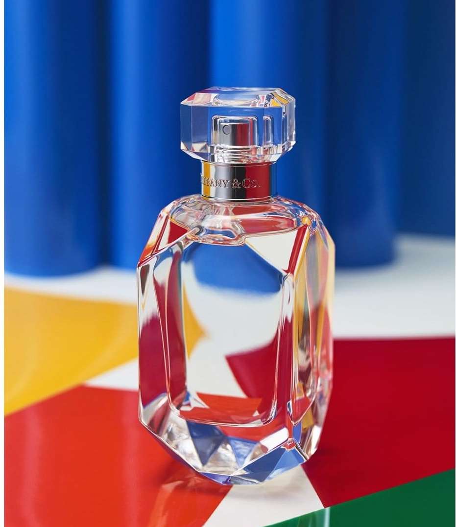 Crystal perfume bottle by Tiffany & Co. with geometric faceted design on a colorful geometric backdrop and blue studio background.