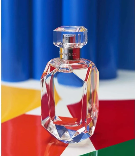 Crystal perfume bottle by Tiffany & Co. with geometric faceted design on a colorful geometric backdrop and blue studio background.