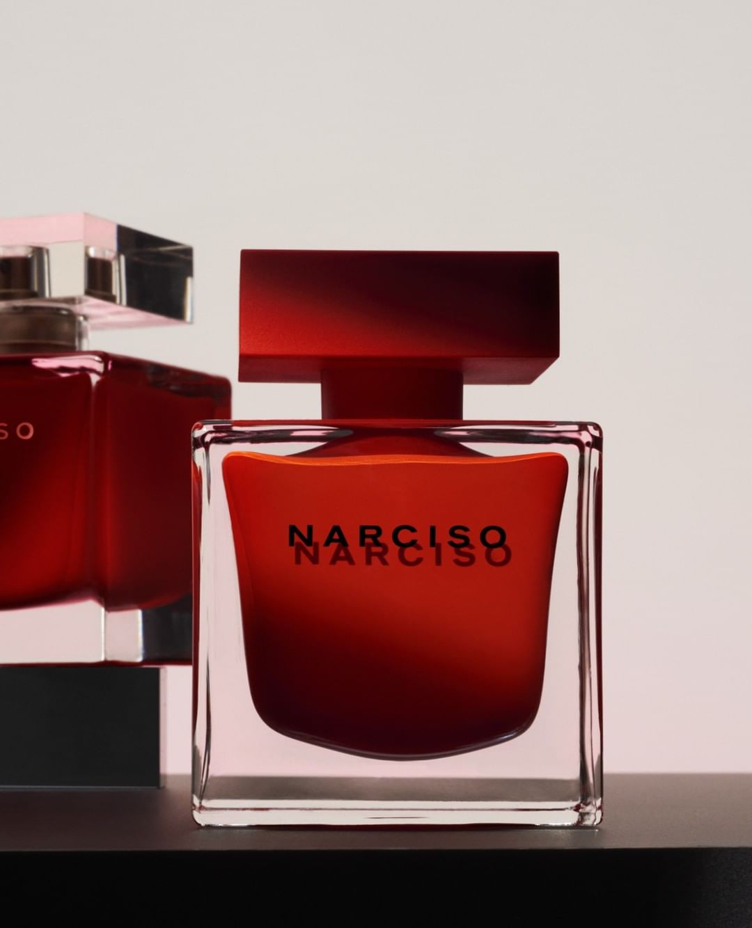 Narciso Rodriguez perfume bottles in square glass containers with red liquid contents on a black surface against a white background.