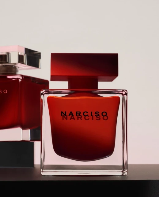 Narciso Rodriguez perfume bottles in square glass containers with red liquid contents on a black surface against a white background.