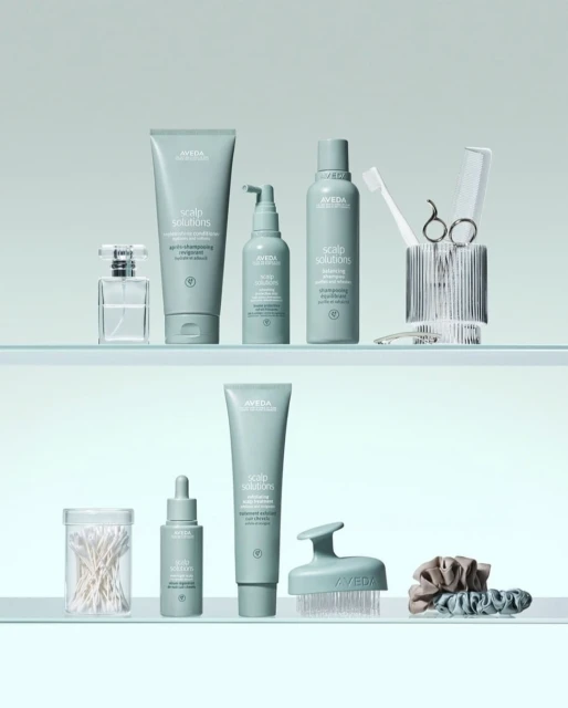 Aveda hair care products arranged on two glass shelves, featuring various scalp solution bottles, brushes, and hair accessories in a clean, minimalist setup.