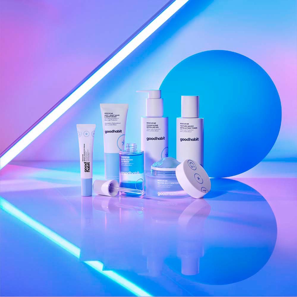 Group of Goodhabit skincare products in sleek white packaging arranged on a reflective surface with neon blue and purple lighting.