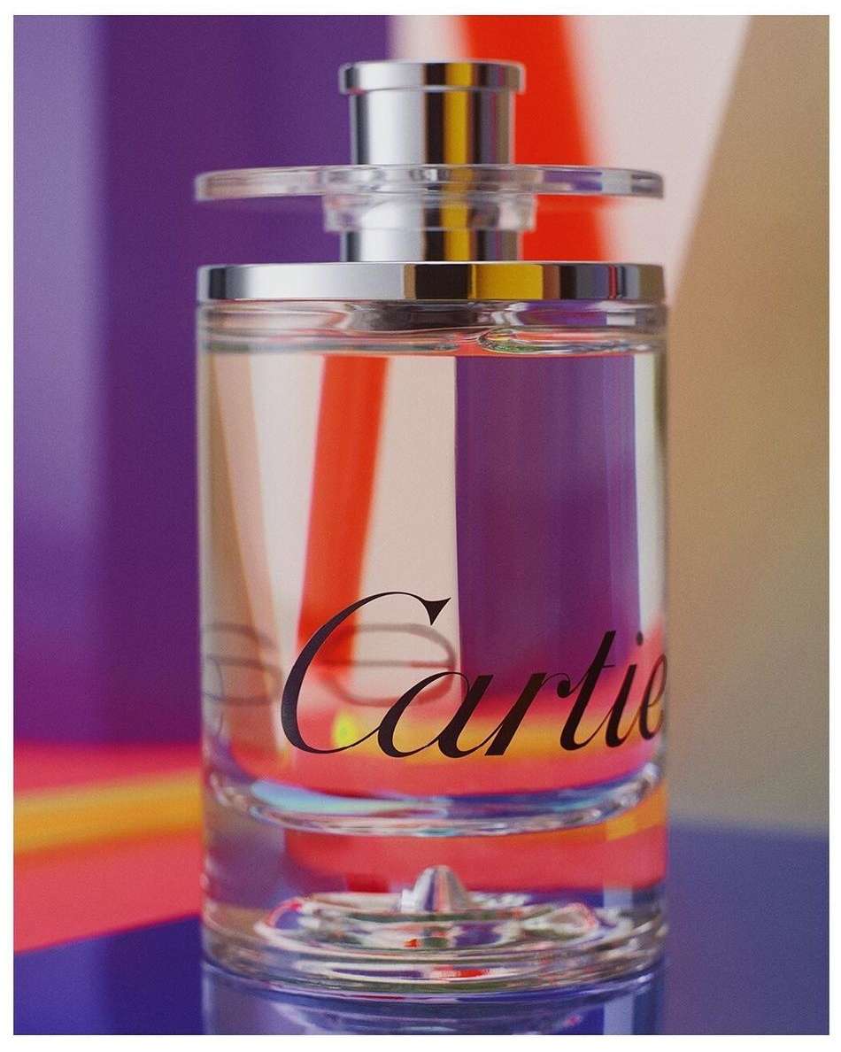 Close-up of a clear Cartier fragrance bottle with a silver cap and black text on a blue table with a purple, orange, and white background.