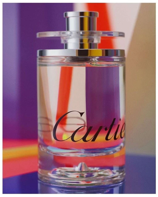 Close-up of a clear Cartier fragrance bottle with a silver cap and black text on a blue table with a purple, orange, and white background.