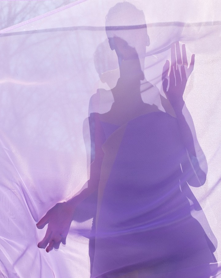 Woman in purple dress visible through a translucent purple fabric, creating a soft, layered effect with diffused lighting and ethereal mood.