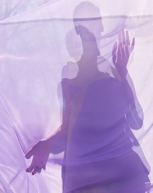 Woman in purple dress visible through a translucent purple fabric, creating a soft, layered effect with diffused lighting and ethereal mood.