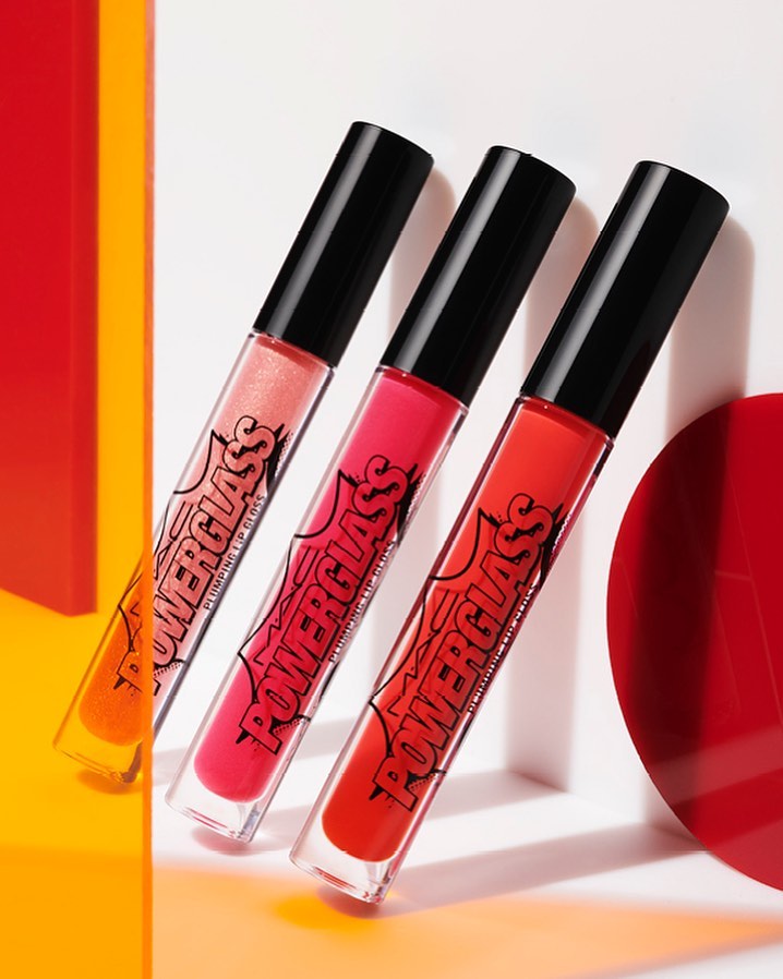 Three tubes of Powerglass Plumping Lip Gloss are diagonally placed against a gradient red and yellow backdrop, capturing a beauty product shot.