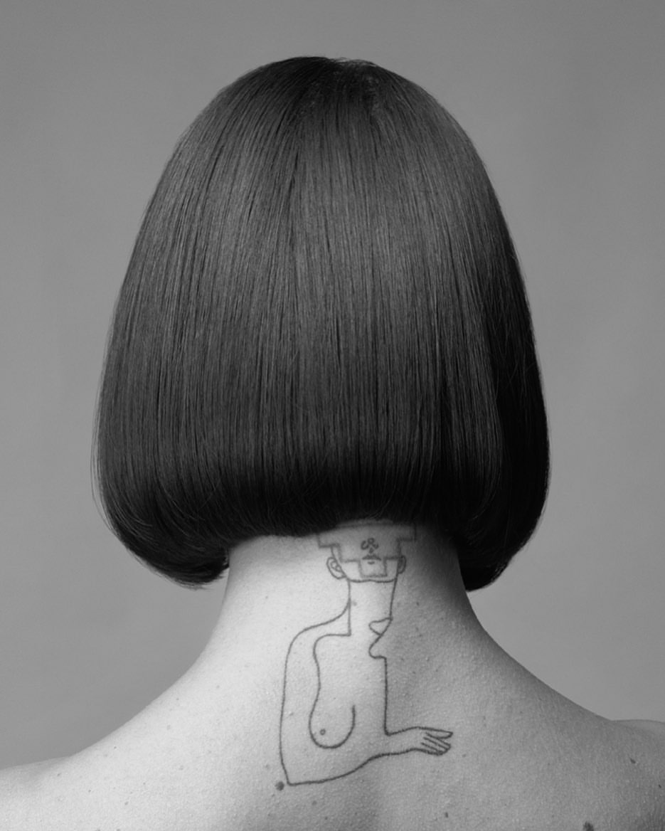Monochrome portrait from behind of a woman with a bob haircut and a line-art tattoo on her neck against a plain gray background.