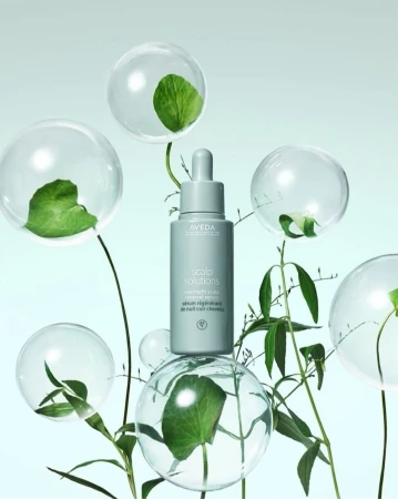Aveda scalp solution bottle with dropper surrounded by clear transparent spheres containing green leaves and stems against a light blue background.