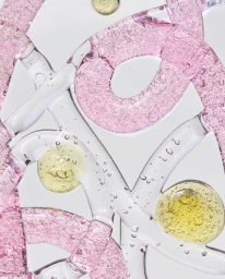 Close-up macro shot of pink and yellow fluid droplets and bubbles trapped within clear glass channels on a white background.