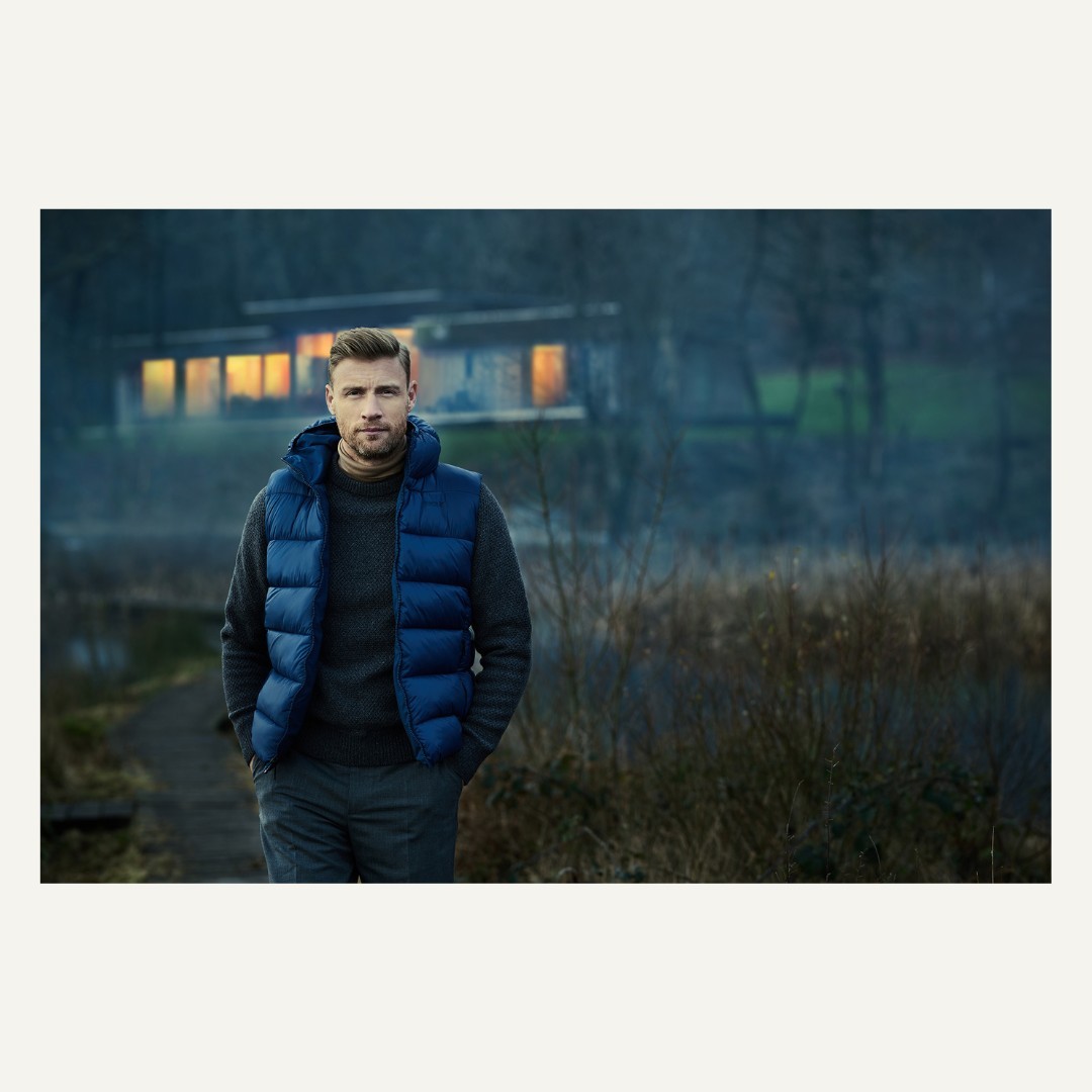 Man in puffer vest walks near modern home with warm lighting. Dark, moody outdoor shot. Wearing sweater and slacks. Casual outdoor fashion.