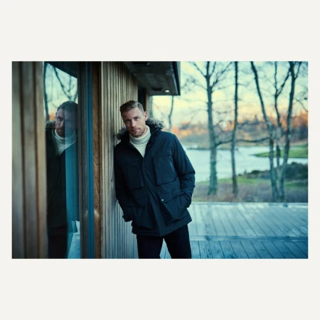 Man in a black winter coat with a fur-trimmed hood leans against a modern wood paneled building with outdoor landscape view and reflection.