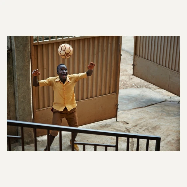 An African boy in a yellow shirt bounces a soccer ball with his head in front of a corrugated metal gate outdoors with concrete.