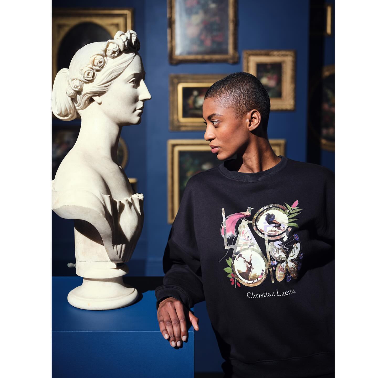Woman in sweatshirt with graphic stands next to a white bust sculpture in an art gallery with framed paintings on a blue wall.