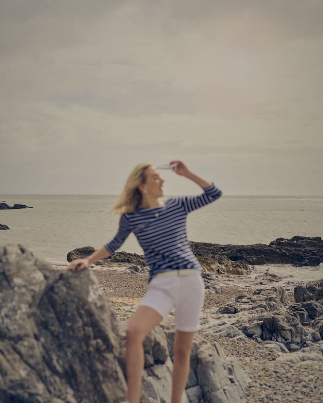 Woman in striped shirt and white shorts stands on rocky shore near ocean, hand shading eyes, looking towards the horizon.