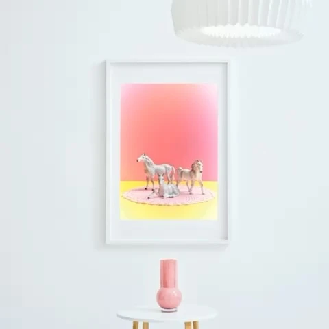 Framed art print of three toy horses on a pink blanket, set against a pink and yellow gradient backdrop, hanging on a white wall.