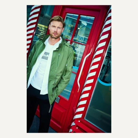 Adult male poses next to a red barber shop doorway with white stripes, wearing a white t-shirt, green jacket, and black pants.