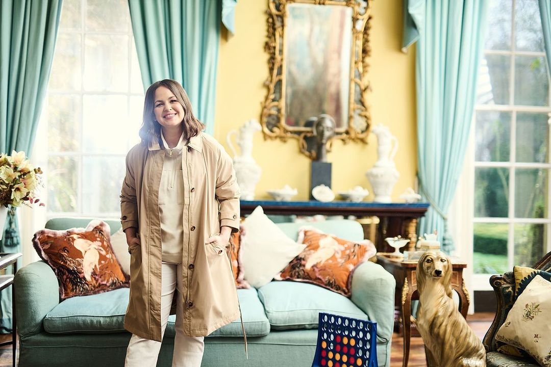 Woman in a beige coat poses in a bright room with a sofa, game, and dog statue. Interior design elements and natural light fill the scene.
