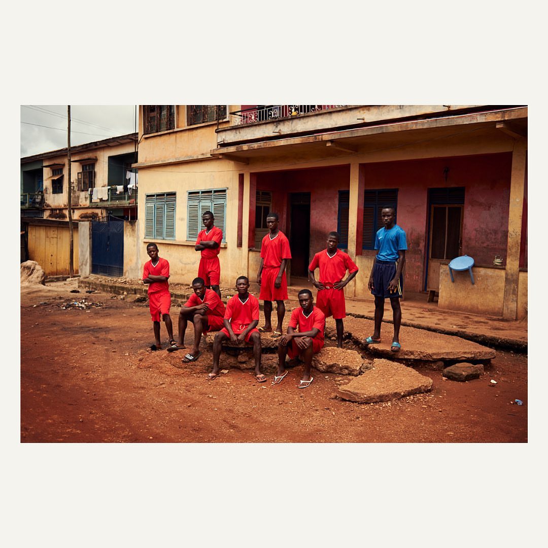 Group of young African men in athletic wear pose outdoors near a building, showcasing a mix of standing and seated poses in a daytime shot.