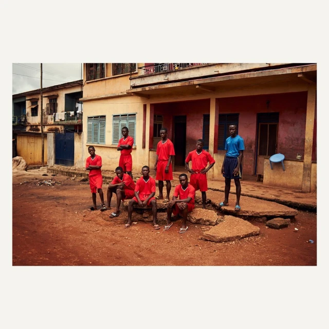 Group of young African men in athletic wear pose outdoors near a building, showcasing a mix of standing and seated poses in a daytime shot.