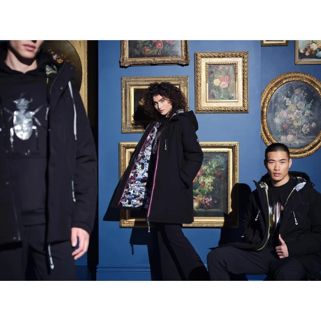 Three people wearing black coats pose in a room with blue walls and framed paintings. Fashion editorial, studio shot, portrait orientation.