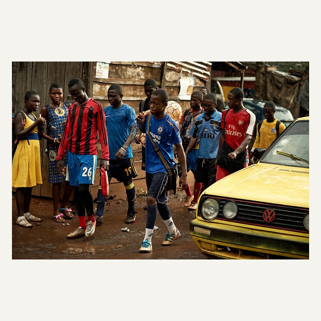 Group of African youth in soccer jerseys walk along a dirt street beside a yellow car in an urban setting, capturing everyday life.