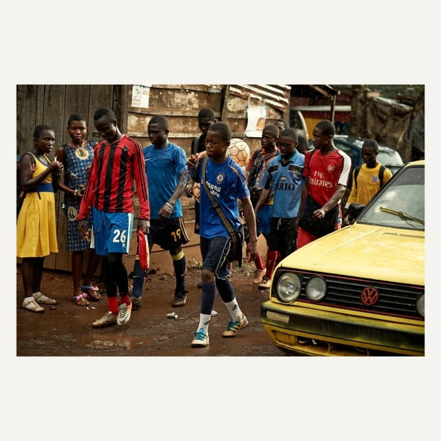 Group of African youth in soccer jerseys walk along a dirt street beside a yellow car in an urban setting, capturing everyday life.