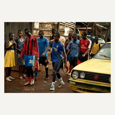 Group of African youth in soccer jerseys walk along a dirt street beside a yellow car in an urban setting, capturing everyday life.