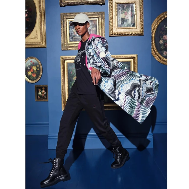 Fashionable woman in patterned coat, black shirt and pants, and boots, posing against a blue wall with framed artwork.
