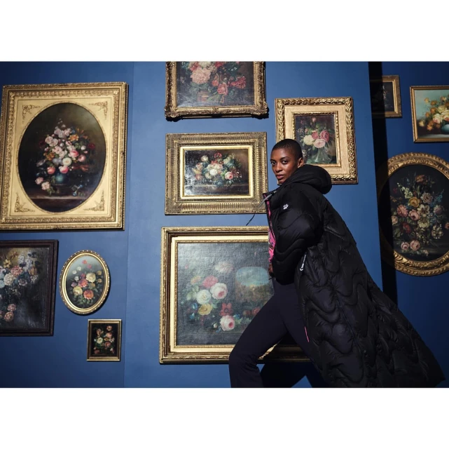 Woman in a long black puffer coat stands in front of a wall adorned with various framed floral paintings in a museum setting.