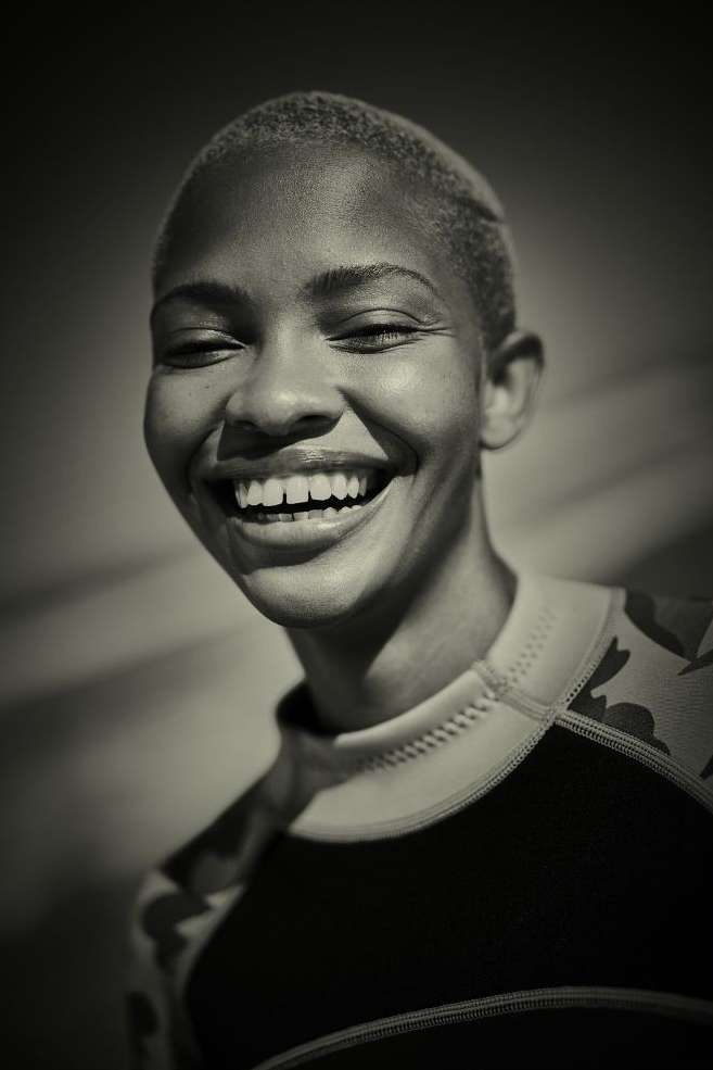 Monochromatic close-up of a smiling adult female with a buzzcut, wearing a patterned rash guard, in front of a neutral background.